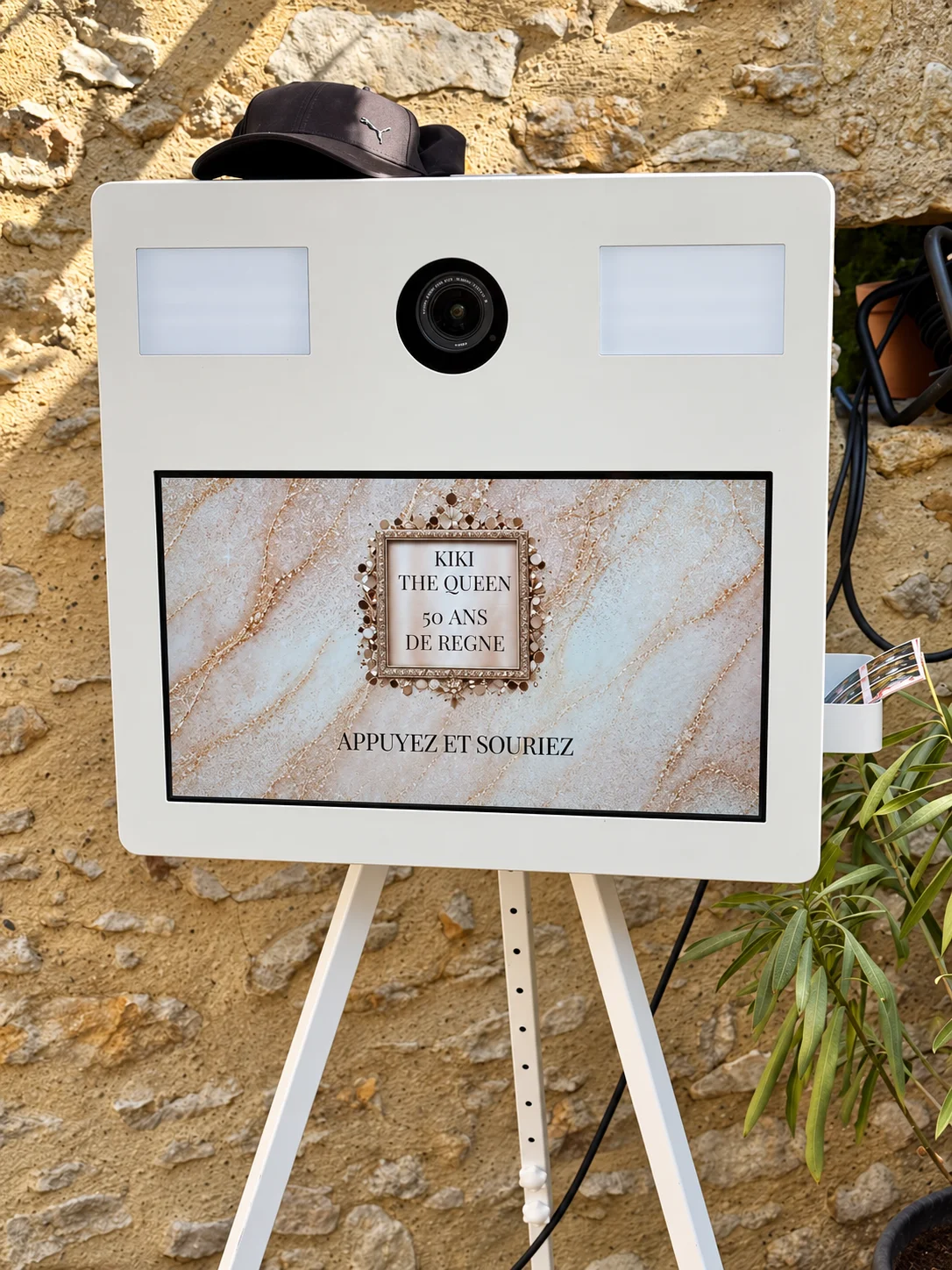 Weiße Fotokabine von Vercors Booth auf Stativ, mit Leinwand, zentraler Linse und zwei Lichtpaneelen, vor einer Steinwand