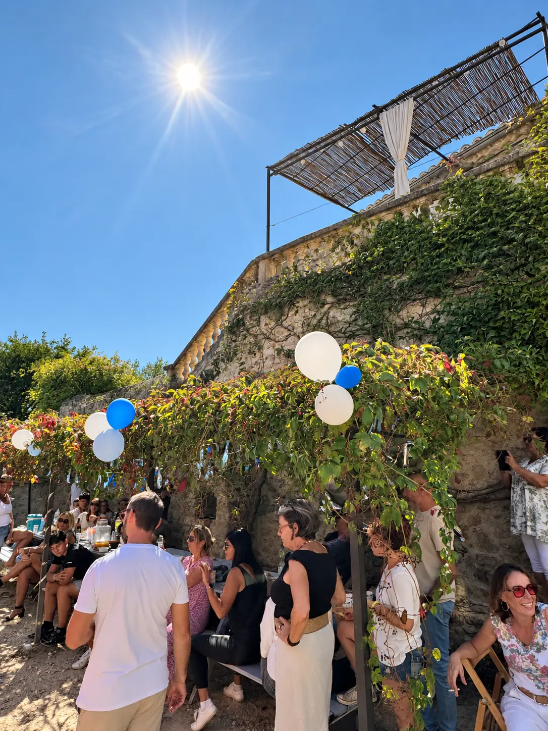 Des proches réunis le long de la façade en pierre, sous les ballons et la vigne.