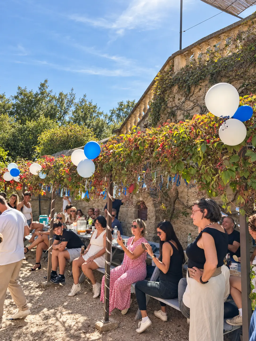 Des invités assis dans la cour, sous une vigne décorée de ballons bleus et blancs.
